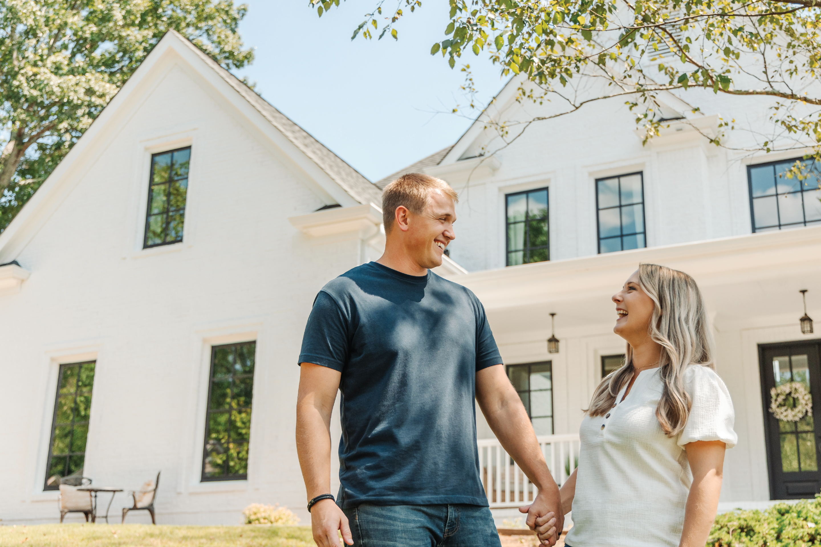 A smiling couple holding hands outside a modern white brick home with large black-framed windows installed by Windows of Wisconsin